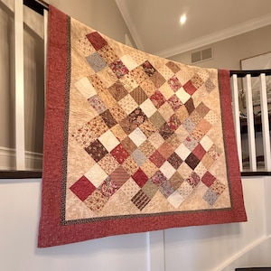 May include: A handmade quilt with a patchwork design, featuring squares in shades of red, brown, beige, and cream. The quilt has a red border and is displayed on a wall. The quilt's pattern includes floral and geometric designs.