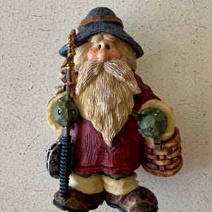 May include: A figurine of a bearded figure wearing a dark blue hat, a red tunic, and light-coloured trousers. The figure holds a tall, textured staff in the right hand and carries a small, brown woven basket in the left hand, all with a carved wood appearance.
