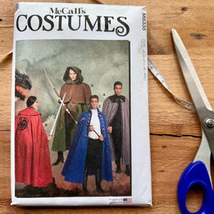 May include: A McCall's Costumes sewing pattern envelope labeled M8335, featuring illustrations of four individuals wearing capes and cloaks in red, green, blue, and gray. The envelope includes size range S-XXL and is positioned next to metal scissors.