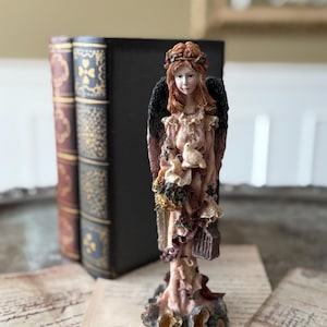 May include: An angel figurine with red hair, black wings, and a pink dress. The angel holds a bird and a small cage. Behind the angel are three antique books with decorative covers. The figurine is on a table with vintage papers.