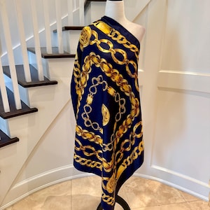May include: A navy blue scarf featuring a repeating pattern of gold-coloured chains and circular medallions. The scarf is draped over a mannequin, displaying a smooth, satin-like texture with a vibrant contrast between the dark blue and metallic gold.