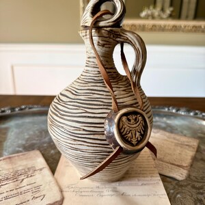 Puede incluir: Una jarra de cerámica con esmalte color crema y líneas horizontales texturizadas en marrón oscuro. La jarra cuenta con una sola asa y un cordón de cuero atado alrededor del cuello, que sostiene un medallón circular con un águila bicéfala en relieve.