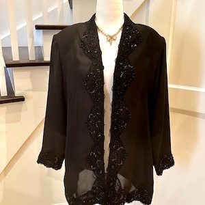 May include: A black sheer jacket with intricate sequin detailing along the lapels, front edges, and cuffs. The jacket is displayed on a mannequin, showcasing its flowing design and elegant embellishments. A gold-toned necklace with a bow pendant is visible.