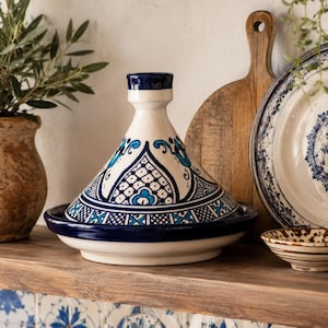 May include: A ceramic tagine featuring a conical lid with a white base and intricate hand-painted patterns in deep blue and turquoise. The base of the tagine is a solid dark blue, and the lid is topped with a small, cylindrical handle.