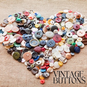 Può includere: Un cuore composto da una varietà di bottoni vintage in vari colori, tra cui rosso, blu, verde e bianco. I bottoni sono disposti su un tessuto strutturato color beige. Le parole "VINTAGE BUTTONS" sono stampate in nero.
