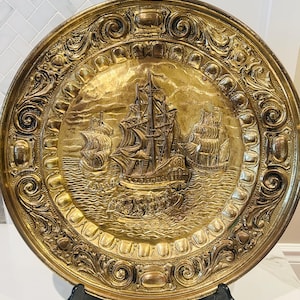 May include: A round, gold-colored metal decorative plate with an embossed nautical scene featuring sailing ships. The plate has an ornate, detailed border with scrollwork and other decorative elements. The plate is displayed on a black stand.