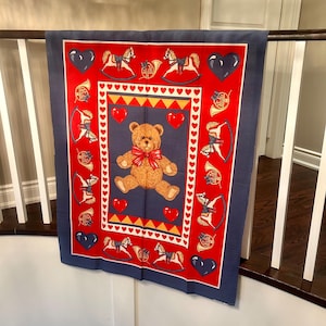 Può includere: Una coperta per bambini rossa e blu navy con un orsacchiotto, cavalli a dondolo, cuori e strumenti musicali. L'orsacchiotto ha un papillon rosso. Il bordo è blu navy.
