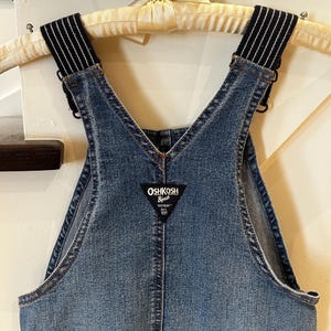 May include: Close-up of a denim overall. The bib overall has adjustable straps with black and white striped elastic. The brand name "OSHKOSH B'gosh" is on a black and white label. The overall is hanging on a wooden hanger.