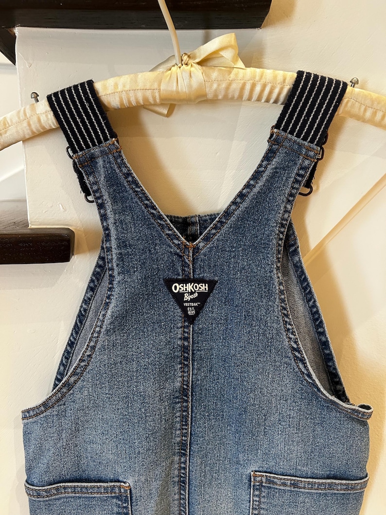 May include: Close-up of a denim overall. The bib overall has adjustable straps with black and white striped elastic. The brand name "OSHKOSH B'gosh" is on a black and white label. The overall is hanging on a wooden hanger.