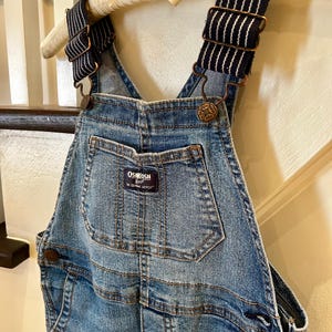 May include: Blue denim overalls with adjustable striped straps and metal hardware. The overalls feature a front pocket and the "OshKosh B'gosh" brand label. The garment is hanging on a wooden hanger.
