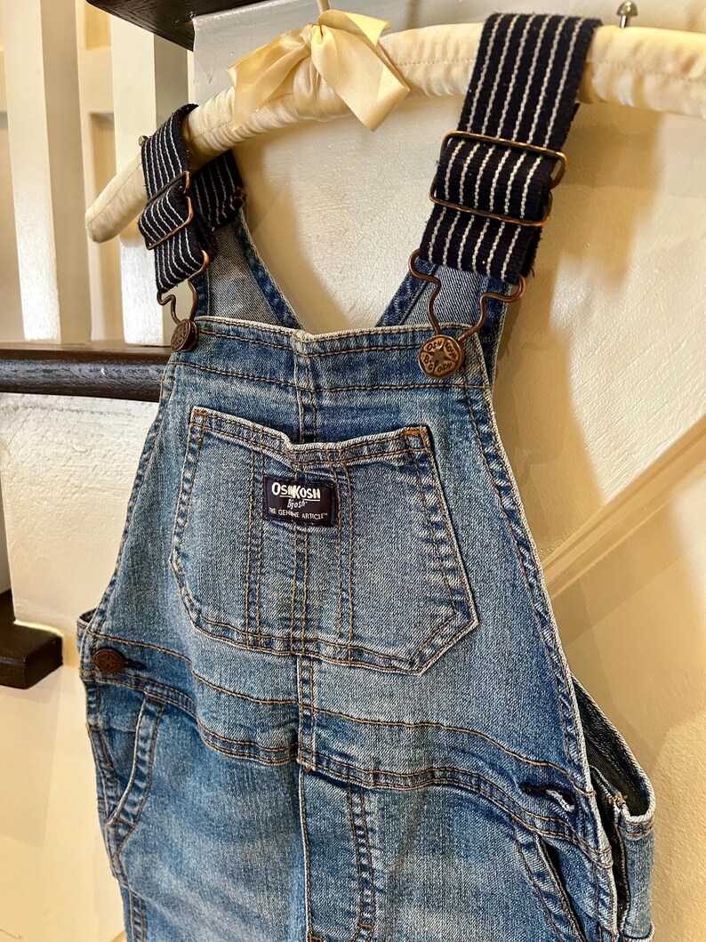 May include: Blue denim overalls with adjustable striped straps and metal hardware. The overalls feature a front pocket and the "OshKosh B'gosh" brand label. The garment is hanging on a wooden hanger.