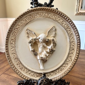 May include: A white ceramic plaque featuring a 3D sculpted angel holding a wreath of flowers. The plaque is framed in an ornate gold and brown metal frame with a decorative stand.