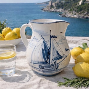Può includere: Brocca in ceramica bianca con un disegno di barca a vela blu. La brocca ha un manico curvo e un beccuccio largo. Sono visibili anche limoni e un bicchiere d'acqua con una fetta di limone. La brocca è su una tovaglia di lino, con il mare sullo sfondo.