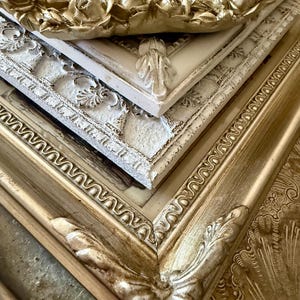 May include: A stack of ornate picture frames in various shades of gold and white. The top frame features a rose design, while others have detailed scrollwork and leaf accents. The frames are arranged at an angle, showcasing their intricate details.