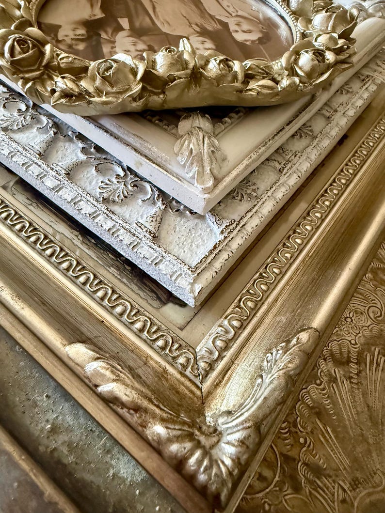 May include: A stack of ornate picture frames in various shades of gold and white. The top frame features a rose design, while others have detailed scrollwork and leaf accents. The frames are arranged at an angle, showcasing their intricate details.