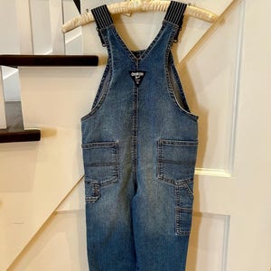 May include: Blue denim overalls with black and white striped straps, hanging on a wooden hanger. The overalls have multiple pockets and the OshKosh logo on the bib. A classic children's garment.