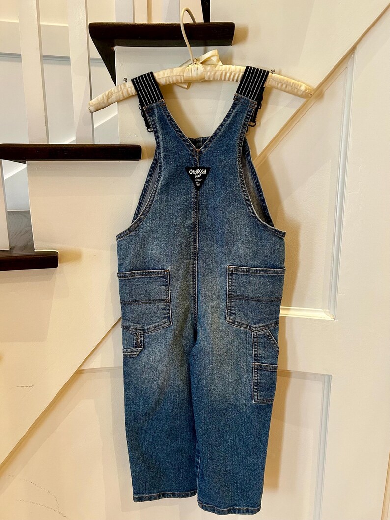 May include: Blue denim overalls with black and white striped straps, hanging on a wooden hanger. The overalls have multiple pockets and the OshKosh logo on the bib. A classic children's garment.