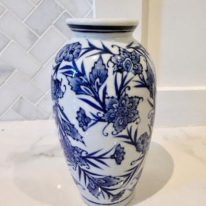 May include: A white ceramic vase featuring a blue floral pattern with intricate leaves and blossoms. The vase has a rounded body, a narrow neck, and a flared rim with a thin blue band around the top edge, showcasing a classic blue and white design.