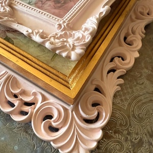 May include: Close-up of two ornate picture frames. The frames are a soft pink color with intricate scrollwork and floral details. The inner frame is gold, and the background features a floral design.