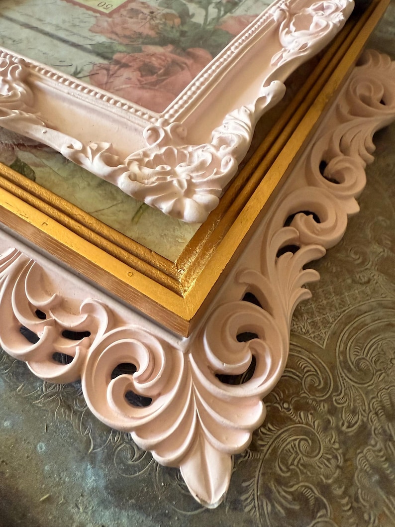 May include: Close-up of two ornate picture frames. The frames are a soft pink color with intricate scrollwork and floral details. The inner frame is gold, and the background features a floral design.
