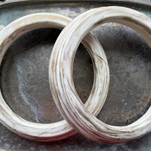 Puede incluir: Dos objetos circulares en forma de anillo con un acabado blanco desgastado y vetas de madera marrón visibles. Los anillos tienen un perfil redondeado y liso, y están colocados superpuestos, mostrando una apariencia texturizada y envejecida.