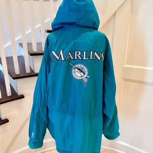 May include: A teal hooded windbreaker jacket featuring the word MARLINS in white lettering across the back. Below the text is a circular Florida Marlins baseball team logo in white, teal, and black. The jacket has long sleeves with elastic cuffs.