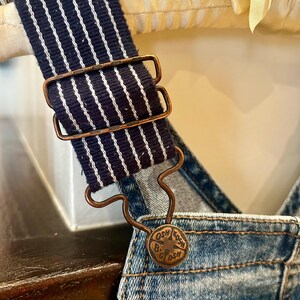 May include: Close-up of denim overalls with a navy blue and white striped strap. The strap is held in place by a copper-colored metal buckle. A copper-colored button with the text "OSH B'GOSH" is visible.