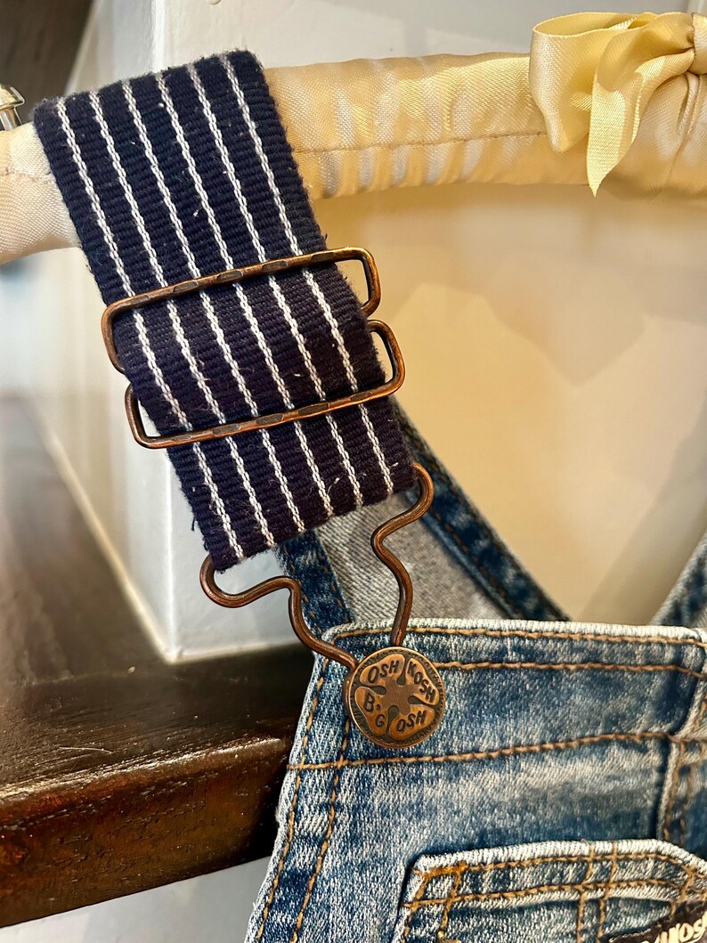 May include: Close-up of denim overalls with a navy blue and white striped strap. The strap is held in place by a copper-colored metal buckle. A copper-colored button with the text "OSH B'GOSH" is visible.