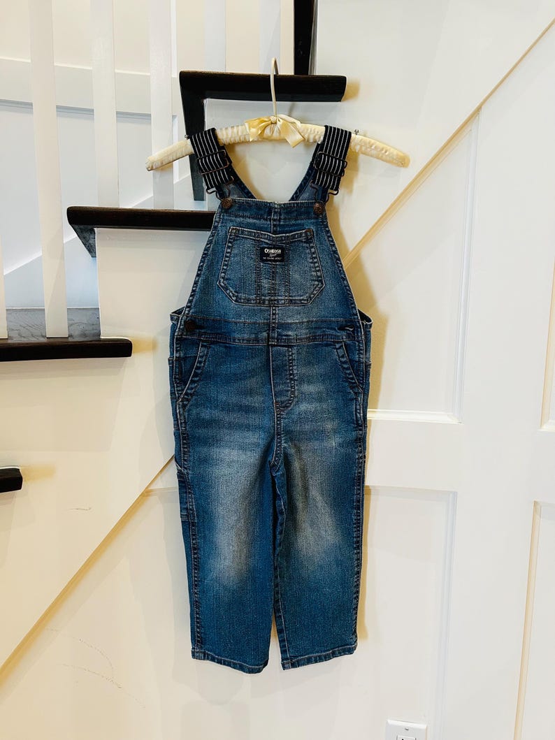 May include: Children's blue denim dungarees with a bib pocket and adjustable straps. The dungarees have a classic design with a faded wash and are hanging on a wooden hanger. The brand name "OshKosh" is visible on the bib pocket.