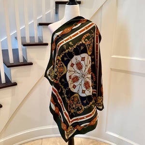 May include: A square scarf featuring a dark green background with a central white circular medallion. The design includes gold, red, and white patterns of belts, buckles, tassels, and ornate filigree motifs printed across the fabric surface.