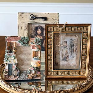 May include: Three decorative picture frames on a gold-colored mirrored tray. The frames feature vintage-style artwork and ornate detailing. One frame has a key attached. The tray sits on a wooden surface.