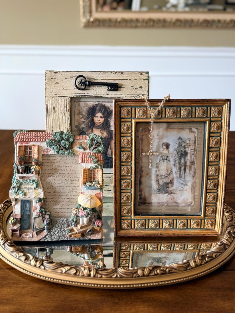 May include: Three decorative picture frames on a gold-colored mirrored tray. The frames feature vintage-style artwork and ornate detailing. One frame has a key attached. The tray sits on a wooden surface.