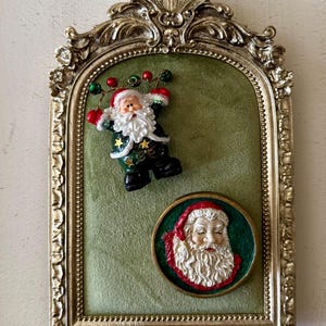 May include: An arched gold-toned ornate frame with a green velvet backing. It features a three-dimensional Santa Claus figure holding a wire with red and green bells, and a circular plaque displaying a relief portrait of Santa Claus's face.
