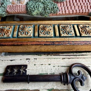 May include: A decorative antique key with a dark brown finish, featuring a detailed handle and a keyhole end with star cutouts. The key rests on a distressed white and green wooden surface, with a gold and teal decorative frame in the background.
