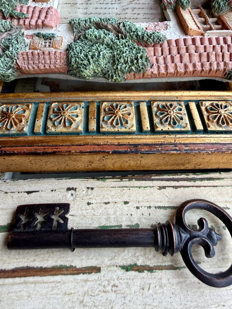 May include: A decorative antique key with a dark brown finish, featuring a detailed handle and a keyhole end with star cutouts. The key rests on a distressed white and green wooden surface, with a gold and teal decorative frame in the background.
