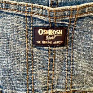 May include: Close-up of denim fabric with visible stitching and a dark blue label. The label reads "OSHKOSH Bgosh THE GENUINE ARTICLE" in white and gold lettering. The denim is a light blue wash.