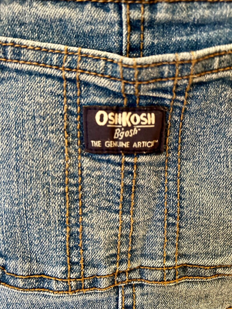 May include: Close-up of denim fabric with visible stitching and a dark blue label. The label reads "OSHKOSH Bgosh THE GENUINE ARTICLE" in white and gold lettering. The denim is a light blue wash.