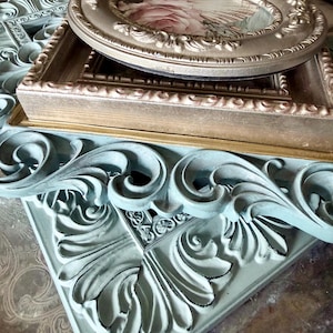 May include: A collection of decorative picture frames in various shapes and sizes. The frames are in shades of gold, beige, and a distressed teal. One oval frame displays a floral print. The frames have ornate detailing, perfect for home decor.