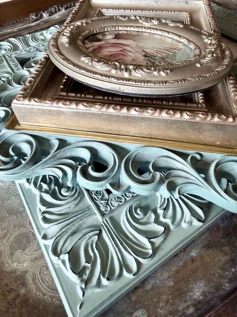 May include: A collection of decorative picture frames in various shapes and sizes. The frames are in shades of gold, beige, and a distressed teal. One oval frame displays a floral print. The frames have ornate detailing, perfect for home decor.