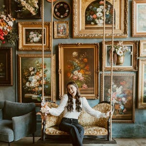 May include: A room featuring a collection of framed floral paintings and decorative plates on a teal wall. A vintage-style swing hangs in front of the wall. A person is sitting on the swing, smiling.
