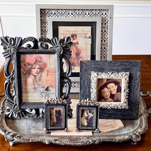 May include: A collection of framed vintage art and photographs displayed on a decorative silver tray. The frames vary in size, shape, and style, including ornate black frames and smaller, detailed metal frames. The images feature portraits and historical scenes.