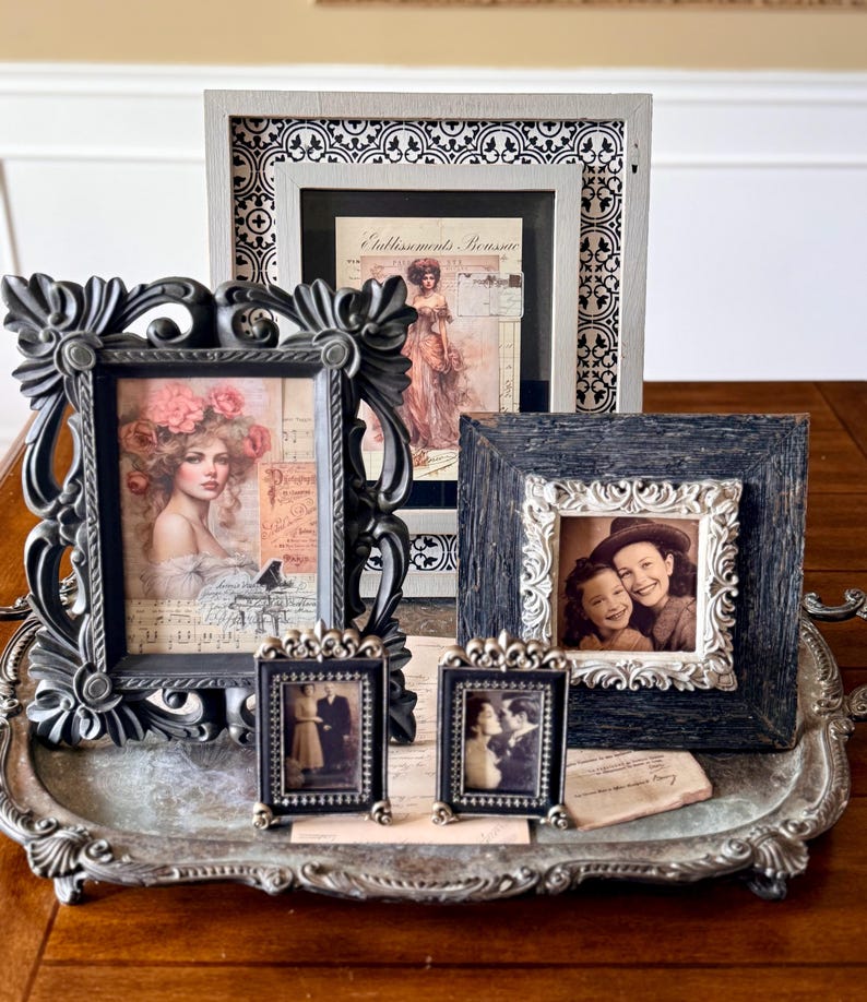 May include: A collection of framed vintage art and photographs displayed on a decorative silver tray. The frames vary in size, shape, and style, including ornate black frames and smaller, detailed metal frames. The images feature portraits and historical scenes.