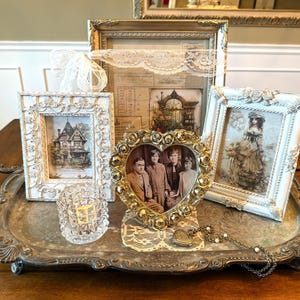 May include: A collection of vintage-style framed pictures and decorative items on a silver tray. The frames are ornate, with images of a house, a portrait, and a woman in a dress. A heart-shaped frame holds a family photo. A small glass candle holder is also present.