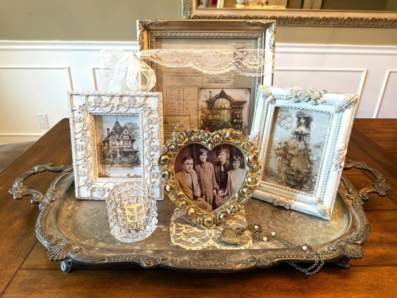May include: A collection of vintage-style framed pictures and decorative items on a silver tray. The frames are ornate, with images of a house, a portrait, and a woman in a dress. A heart-shaped frame holds a family photo. A small glass candle holder is also present.