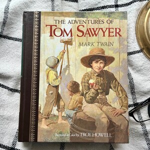 Op de afbeelding: Een vintage hardcover boek getiteld "De avonturen van Tom Sawyer" door Mark Twain. De omslag toont een illustratie van drie jongens, een schildert een hek, een ander zit op een vat en de derde houdt een appel vast. Het boek is geïllustreerd in kleur door Troy Howell.