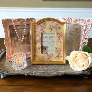 May include: Three decorative picture frames in pink and gold, arranged on a silver tray with a crystal candle holder and a cream-colored rose. The frames feature floral and vintage-style designs, perfect for home decor.