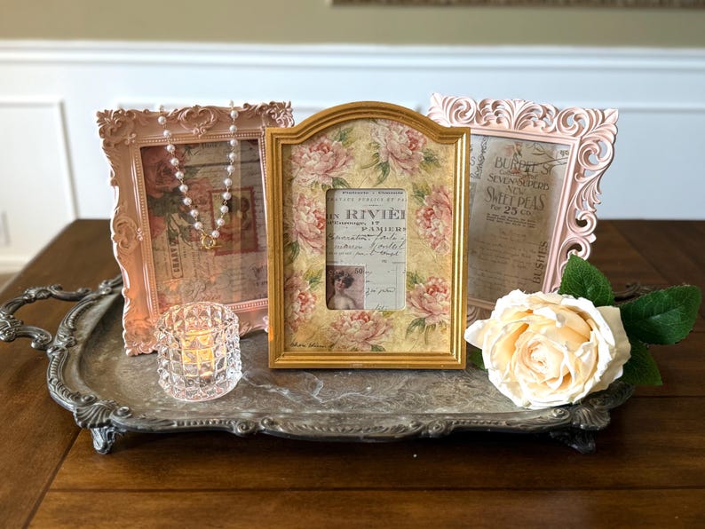 May include: Three decorative picture frames in pink and gold, arranged on a silver tray with a crystal candle holder and a cream-colored rose. The frames feature floral and vintage-style designs, perfect for home decor.