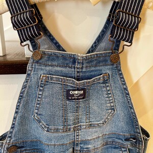 May include: Blue denim overalls with navy and white striped straps. The overalls feature a front pocket with the "OshKosh B'gosh" logo. The straps are attached with bronze-colored metal hardware. The overalls are hanging on a wooden hanger with a cream-colored bow.