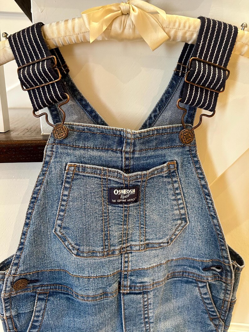 May include: Blue denim overalls with navy and white striped straps. The overalls feature a front pocket with the "OshKosh B'gosh" logo. The straps are attached with bronze-colored metal hardware. The overalls are hanging on a wooden hanger with a cream-colored bow.