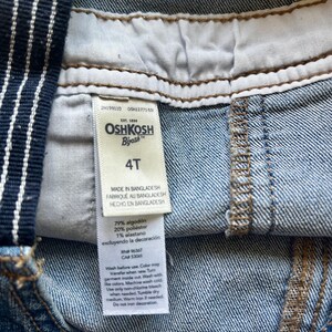 May include: Close-up of a pair of denim overalls with a white and navy blue striped strap. The tag reads "OSHKOSH B'gosh" and "4T". The overalls are made in Bangladesh and are composed of cotton, polyester, and elastane.